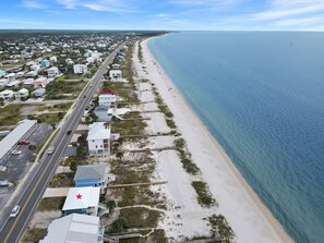 A coastal residential area with houses along a beachfront road, a clear blue sky, and calm sea.