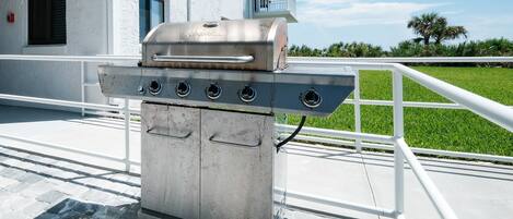 Una parrilla de acero inoxidable con cuatro quemadores, ubicada en un balcón con barandal blanco y vista a un césped verde y un edificio al fondo.