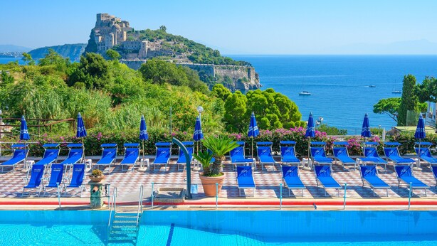 A poolside area with blue lounge chairs and umbrellas, a clear view of the sea, and a historic building on a cliff. - Hotel Parco Cartaromana (Ischia)