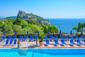A poolside area with blue lounge chairs and umbrellas, a clear view of the sea, and a historic building on a cliff. - Hotel Parco Cartaromana (Ischia)