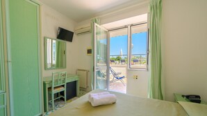 A hotel room with a bed, a desk, a chair, a TV, and a view of the beach through the door. - Hotel Parco Cartaromana (Ischia)