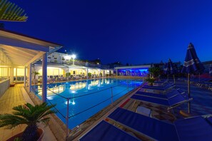 Une piscine entourée de chaises longues et de parasols, elle-même bordée d’une zone couverte aménagée avec des sièges et des plantes en pot.