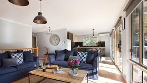 A modern living room with dark blue sofas, a wooden coffee table with books and a vase of flowers, and a kitchen in the background.