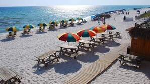 Ein Strand mit Holztischen, bunten Sonnenschirmen, einer Holzterrasse und einem Gebäude mit einem Reetdach.