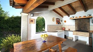 An outdoor kitchen with a brick oven, wooden beams, and a table with a bowl of fruit.