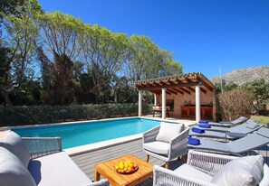 Zona junto a la piscina con sillones de descanso, una mesa de madera con fruta y una estructura de pérgola.