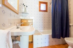 Un baño con un lavamanos blanco, una bañera, y un inodoro. Hay una ventana con marco de madera y una cortina de ducha.
