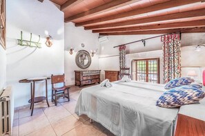 Une chambre à coucher avec un lit, une chaise, une table, une commode et une fenêtre avec des rideaux.