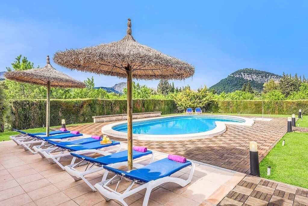 A poolside area with lounge chairs and thatched umbrellas, surrounded by greenery and mountains.