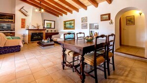 A cozy living room with a fireplace, wooden beams, and a dining area with a table and chairs. - Villa Geroni, Charming 3 bedroomn villa with pool (Pollença)