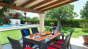 An outdoor dining area with a pool, wicker chairs with red cushions, and a table set for four. - Villa Burgues, Idyllic location affording beautiful views. Alcudia (Alcúdia)
