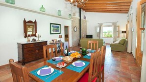 A dining area with a wooden table set for four, surrounded by chairs. There is a wooden cabinet to the left and a green sofa in the background. - Villa Burgues, Idyllic location affording beautiful views. Alcudia (Alcúdia)