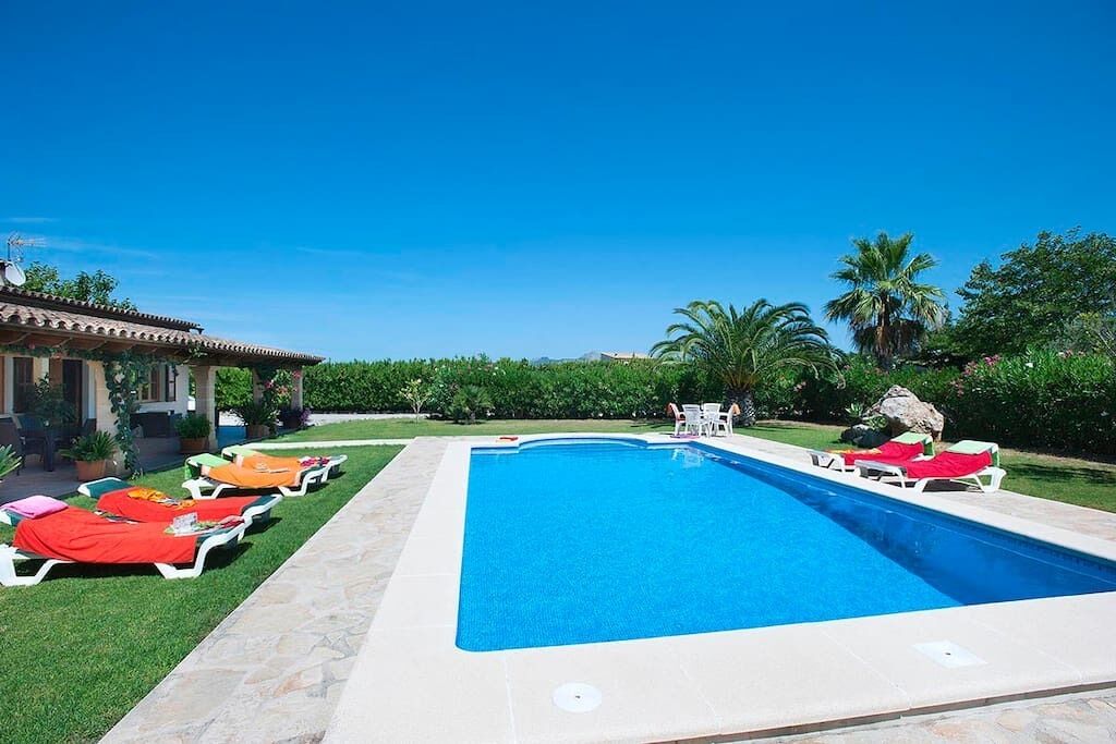 Ein rechteckiger Pool mit glasklarer blauer Wasserfläche, umgeben von einer gefliesten Liegefläche. Entlang des Poolrandes reihen sich Liegestühle mit roten und weißen Kissen.