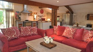 Uma sala de estar aconchegante com um sofá vermelho, uma mesa de centro de madeira com um arranjo floral e uma cozinha ao fundo, com vigas de madeira e um relógio vermelho.