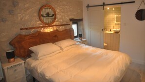 Quarto com parede de pedra, cabeceira de madeira, cama branca, mesa de cabeceira, abajur e banheiro visível por uma porta aberta.