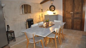 Uma sala de jantar com uma mesa comprida, cadeiras brancas, um banco de madeira e um vaso com planta.