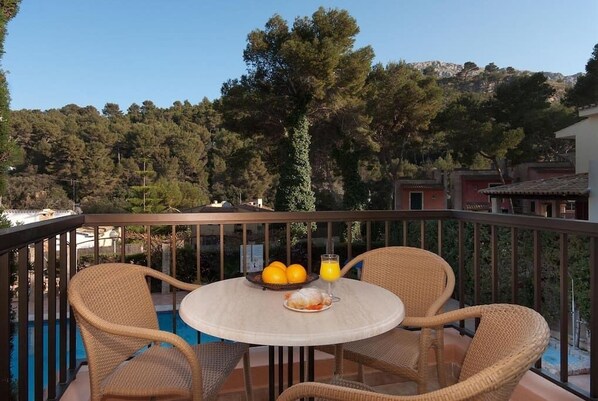 Un balcon avec des fauteuils en osier et une table, une piscine et une vue sur une colline boisée.