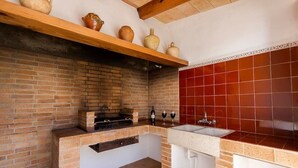 Une cuisine avec un mur en briques, un plan de travail recouvert de carreaux rouges, une étagère en bois ornée de poteries, un évier et un barbecue encastré.