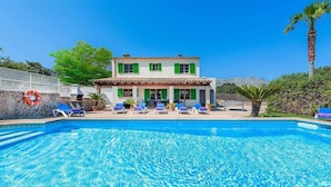 Una casa de dos plantas con piscina, contraventanas verdes y mobiliario de exterior.