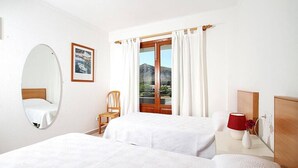 A hotel room with two beds, a wooden headboard, a large mirror, a window with curtains, a chair, a nightstand with a lamp, and a framed picture on the wall. - Villa Animeta, Hermosa villa cerca de Port Pollensa (Pollença)