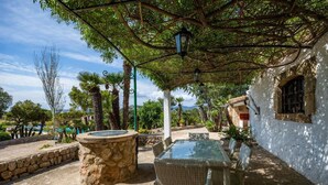 A covered outdoor area with a stone fountain, wicker furniture, and a white building with a window. - Villa Piedra, Surrounded by nature, but close to Port Pollensa (Pollença)