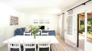 A dining area with a white table and chairs, a blue sofa, and a TV. There are three framed pictures on the wall, a vase with flowers, and a sliding glass door leading to a balcony. - Villa La Plana, Fantastic villa in a very quiet area (Pollença)