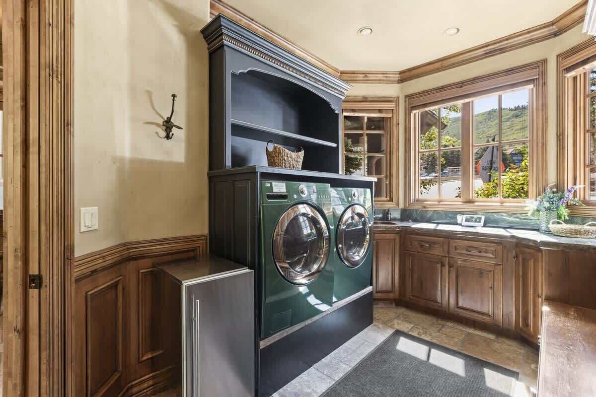 A laundry room with a washing machine, wooden cabinets, a window with a view, and a shelf with a basket.