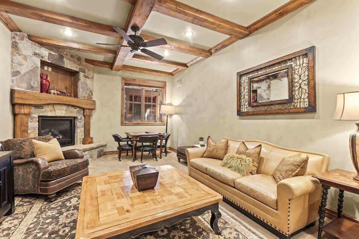 A living room with a stone fireplace, wooden ceiling beams, a ceiling fan, a dining area, and a large mirror on the wall.