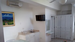 Een kleine, schone kamer met kitchenette, een eethoek met tafel en stoelen, een tv aan de muur en een inloopkast met schuifdeuren.