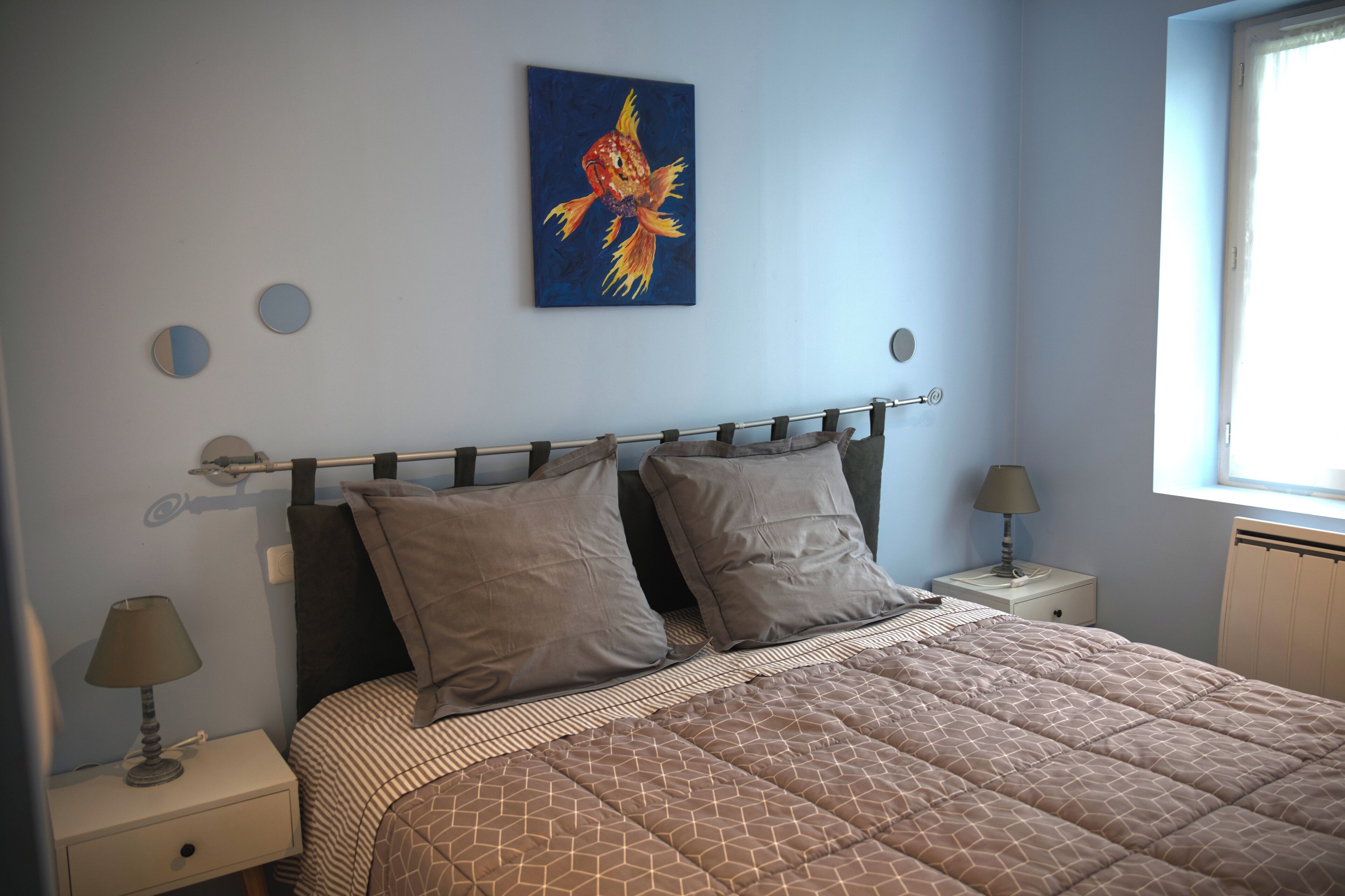 A bedroom with a bed, two pillows, a nightstand with a lamp, a wall clock, and a painting.