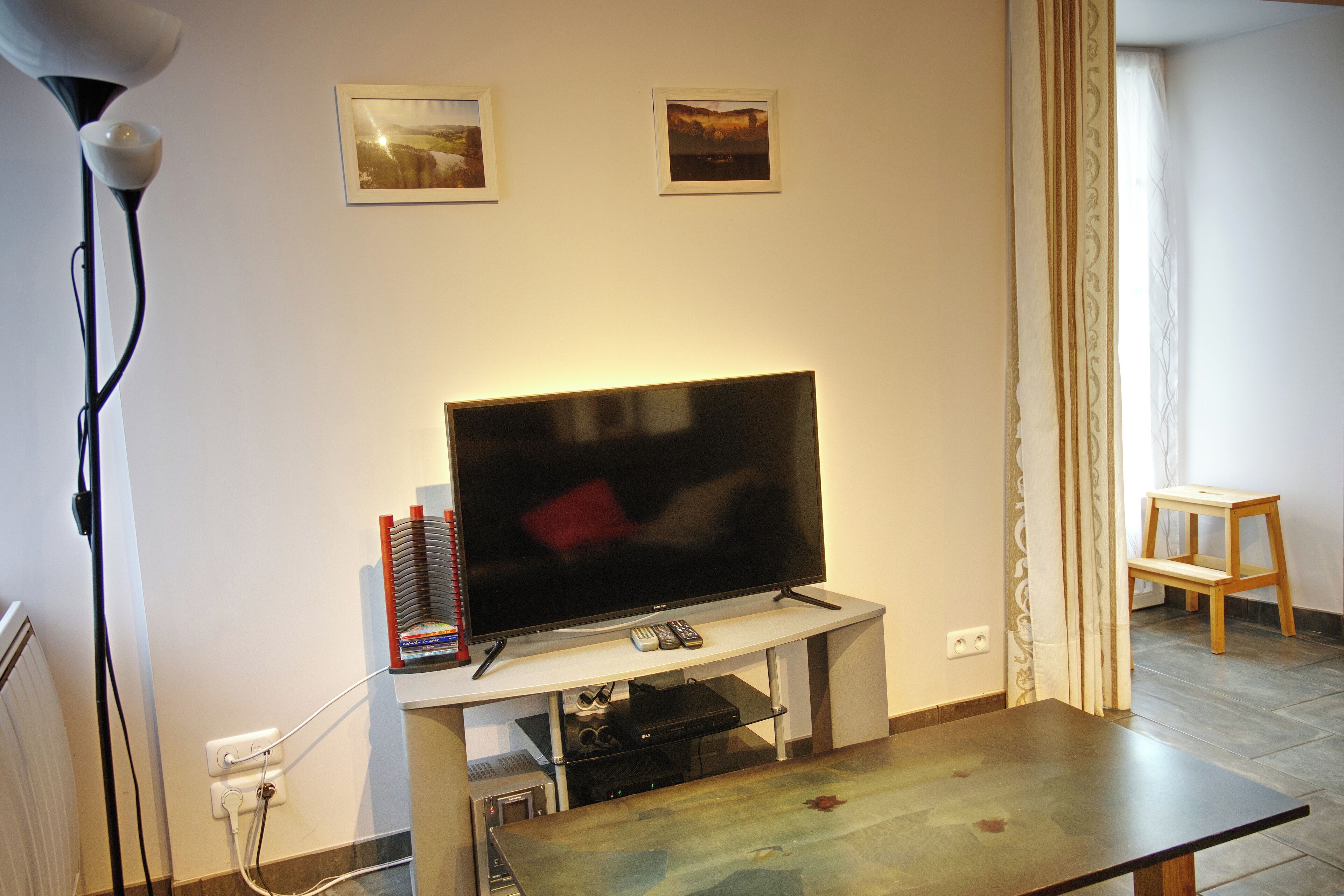 A living room with a flat-screen TV on a stand, a lamp, a small table, and two framed pictures on the wall.