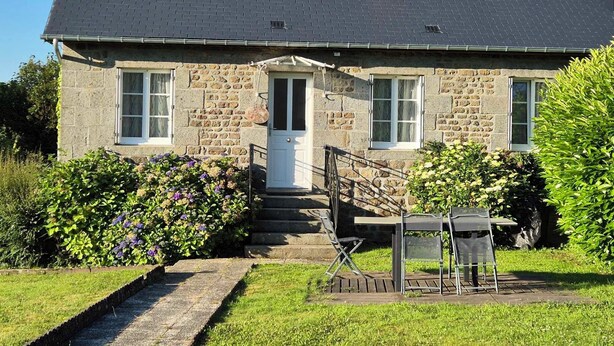 Una casa de piedra con techo gris, una puerta principal y un pequeño porche con mobiliario exterior.