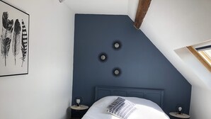 Ein Schlafzimmer mit schrägverdeckter Decke, einem Bett mit blau-weiß gestreifter Bettdecke, zwei Nachttischen mit Lampen und einer wandmontierten runden Leuchte.