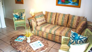 A living room with a striped sofa, a green chair, a round coffee table with magazines and a vase of dried plants, and a painting of an underwater scene on the wall. - Mermaids Retreat Mins to Beach Family Condo (Destin)