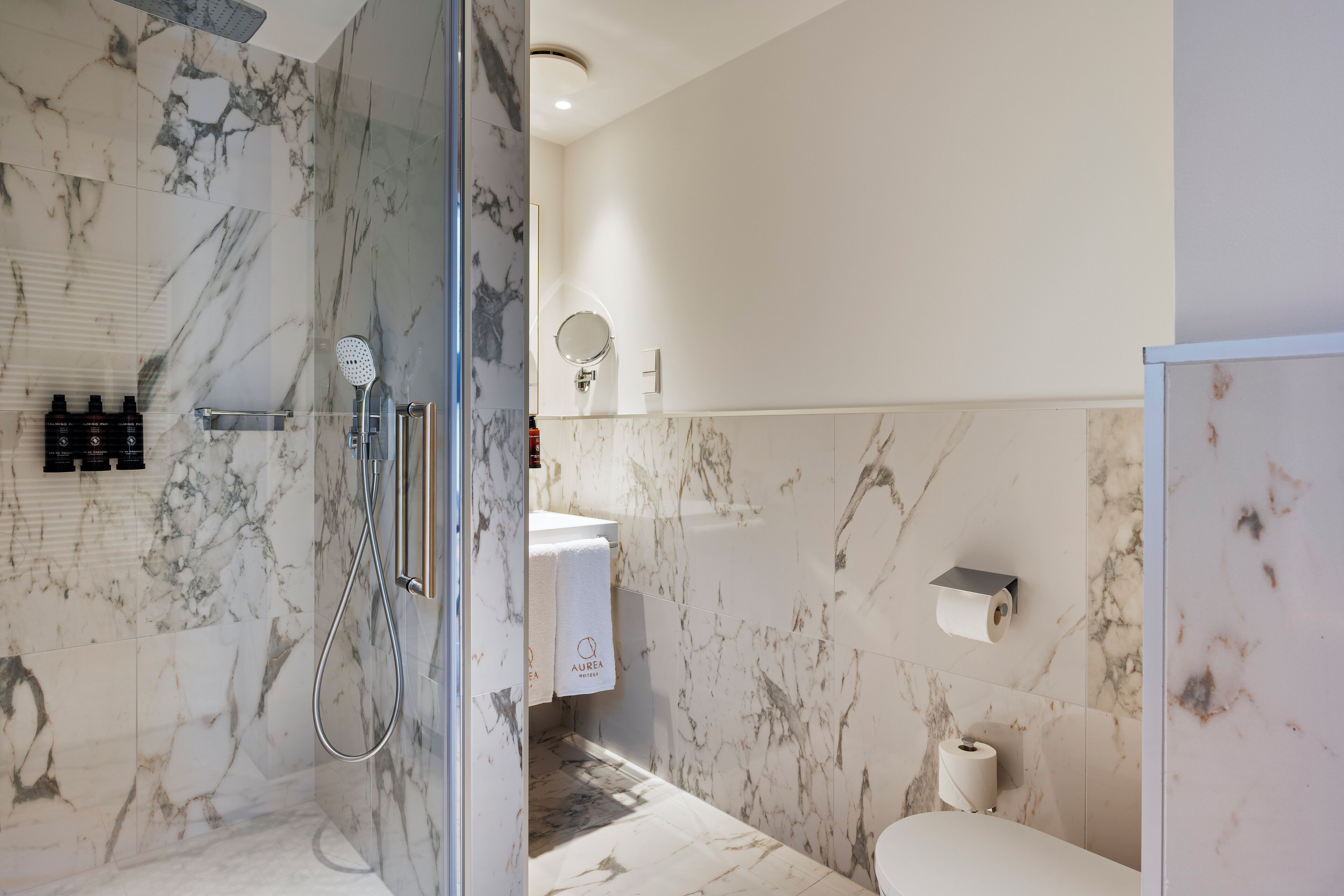 Un baño moderno con ducha de vidrio, paredes de mármol, inodoro y lavamanos con toallero.