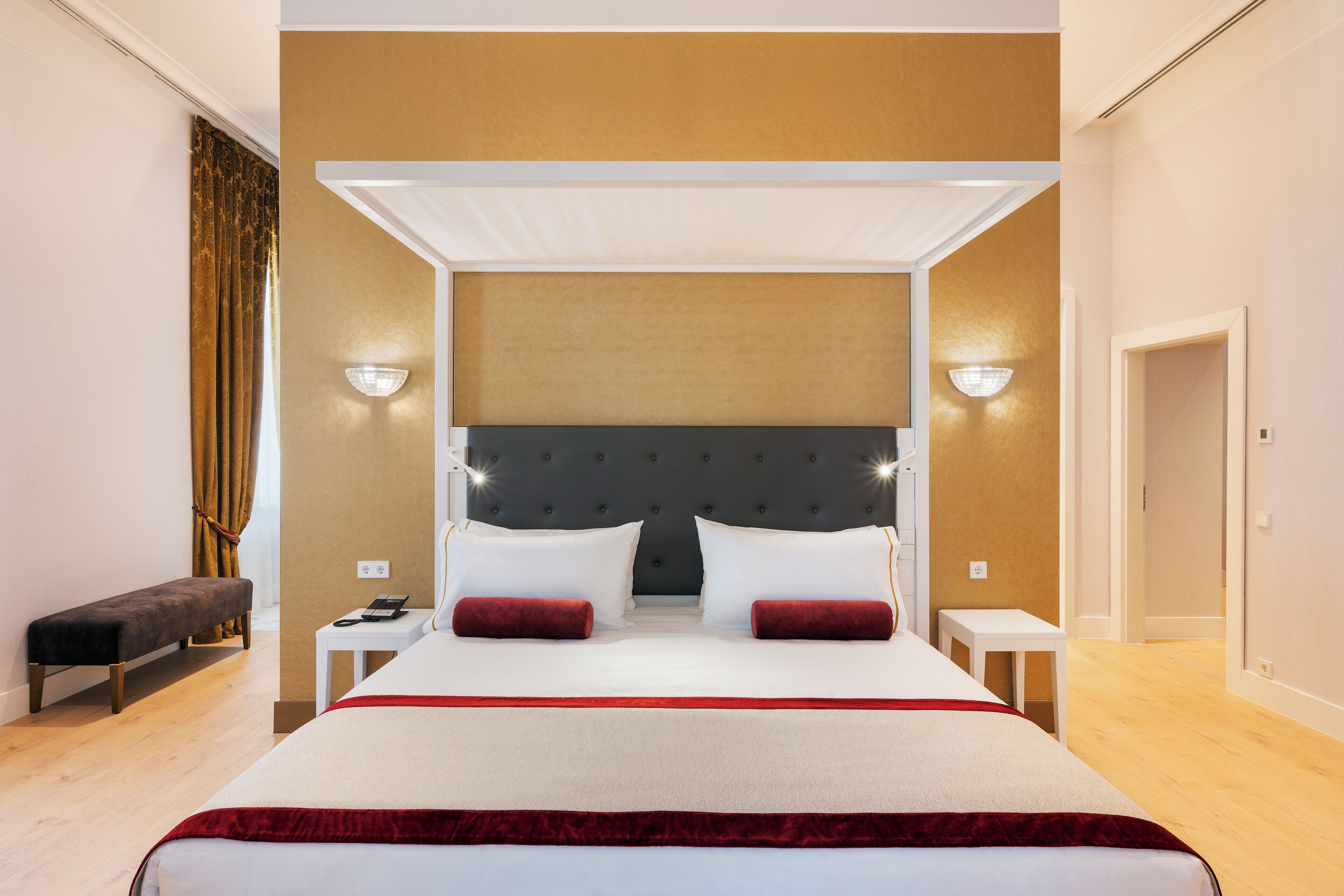 A large bed with a headboard, two bedside tables, a bench, and a wall-mounted light fixture.