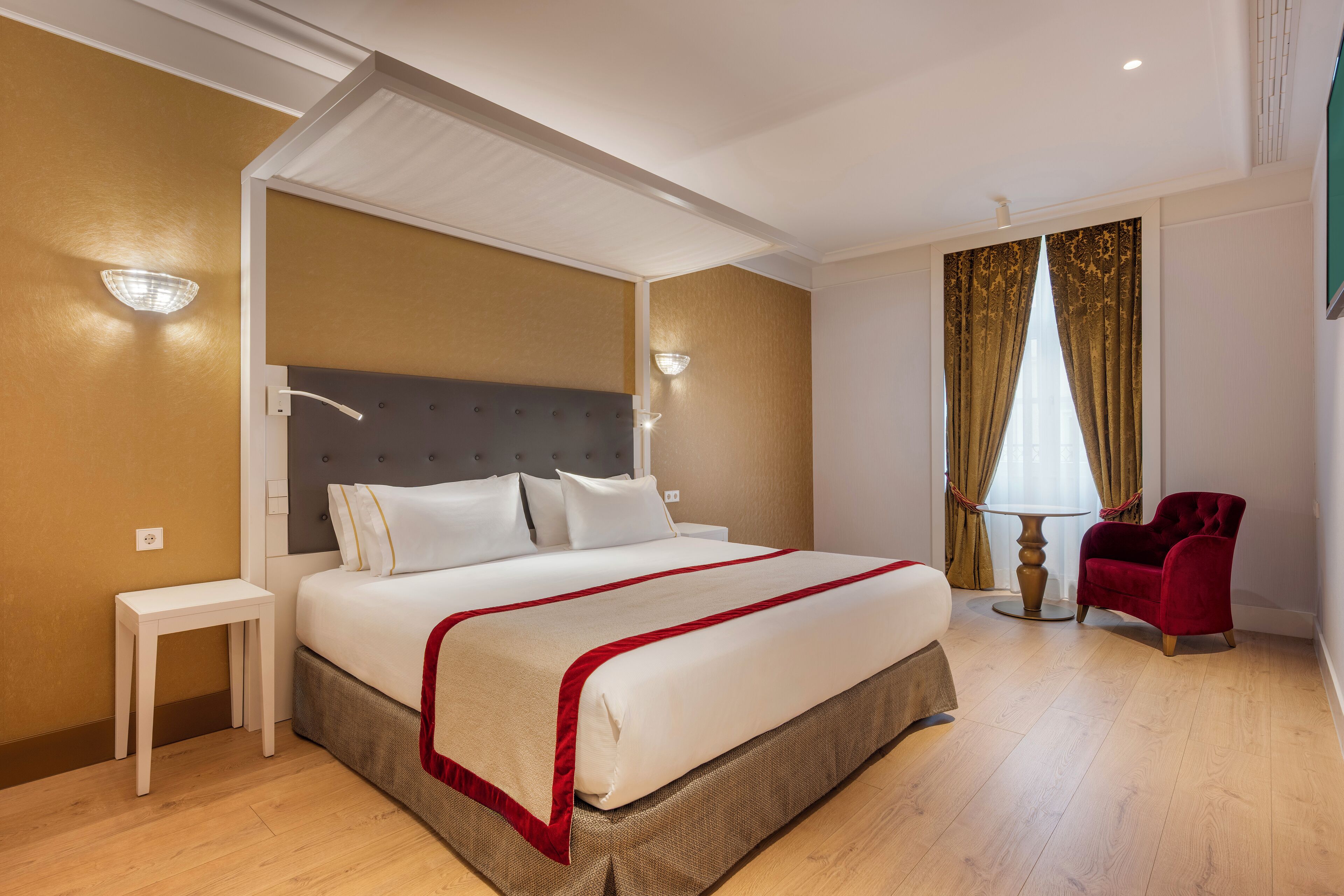 A hotel room with a large bed, a bedside table, a red chair, and a window with curtains.