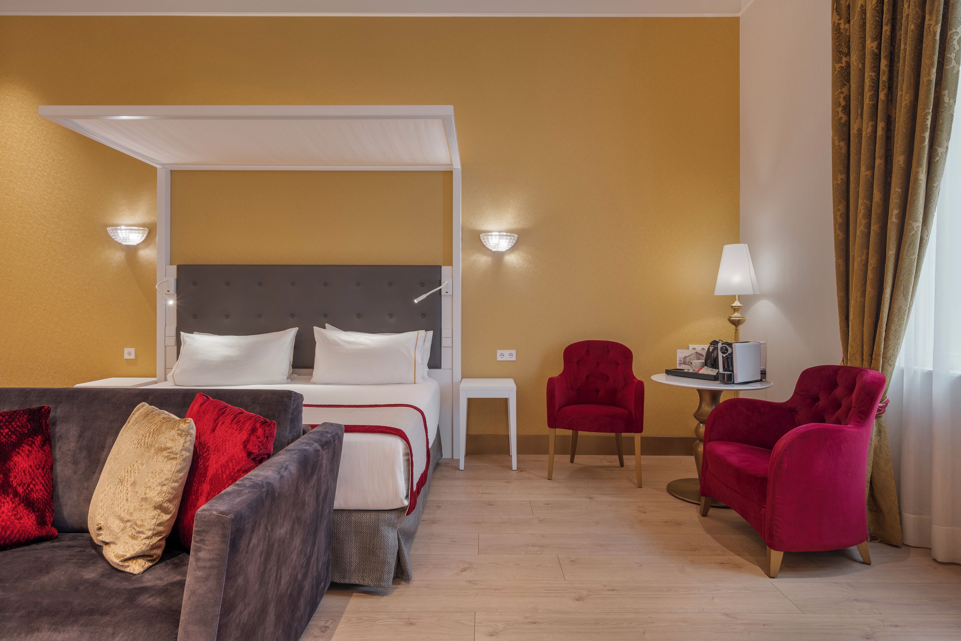 A hotel room with a bed, a sofa, a red armchair, a small table with a lamp, and a wall-mounted light fixture.