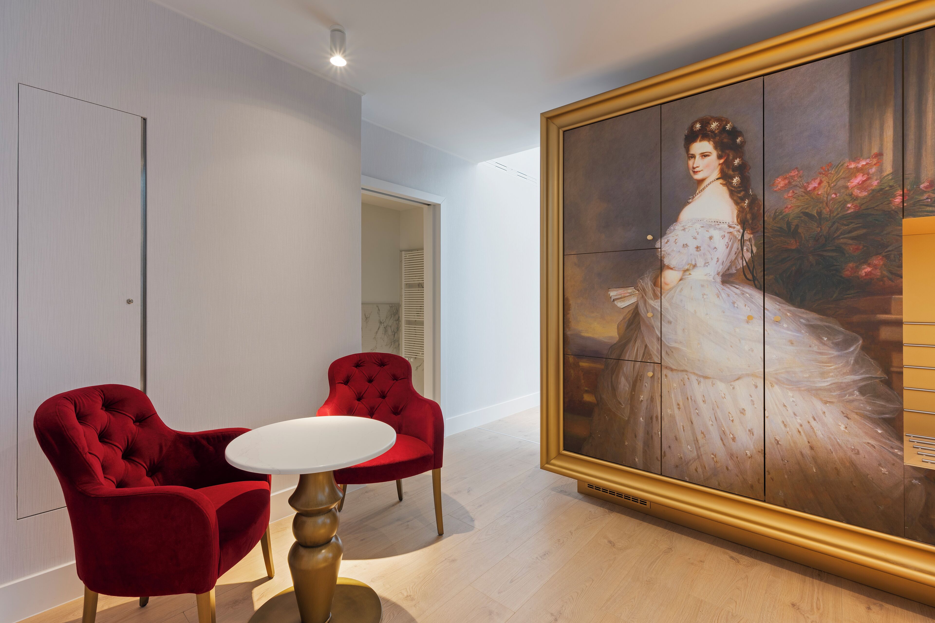 Una habitación con una silla tapizada de rojo, una mesa blanca y un cuadro grande con retrato de una mujer vestida de blanco.