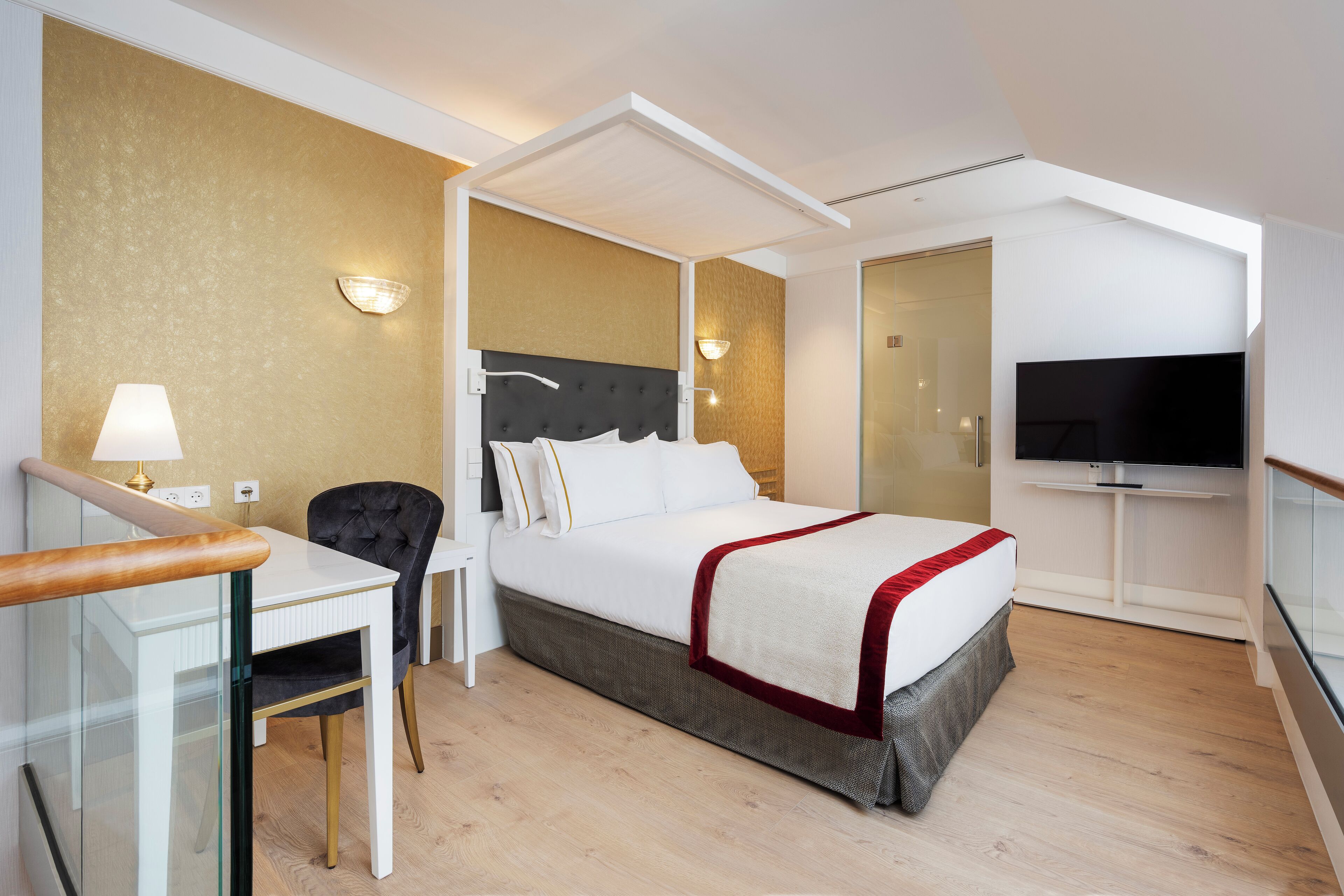 A modern hotel room with a bed, a desk, a chair, a TV, and a glass-enclosed shower area.