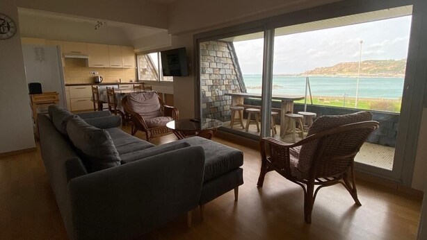 Un salon moderne avec un canapé et des fauteuils en osier, une cuisine à l’arrière-plan et une grande fenêtre donnant sur la mer.