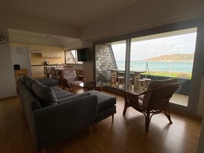 Un salon moderne avec un canapé et des fauteuils en osier, une cuisine à l’arrière-plan et une grande fenêtre donnant sur la mer.