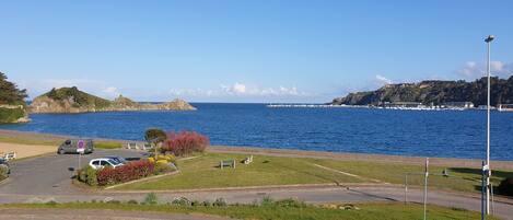A coastal road with a parking area, benches, and a view of the sea and a lighthouse.