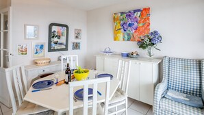 A dining area with a white table, blue plates, and a yellow bowl. There are framed pictures on the wall and a large painting of flowers. - La Lagune - Appartement vue mer (Pléneuf-Val-André)
