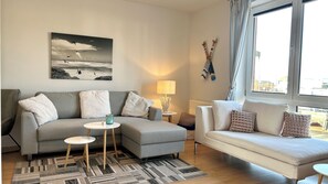 Ein Wohnzimmer mit grauem Sofa, weißem Sofa, einem kleinen runden Tisch, einer Lampe und einem Gemälde von Vögeln über Wasser.