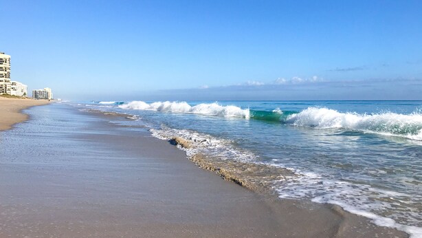 A beach with waves, a clear sky, and buildings in the background.