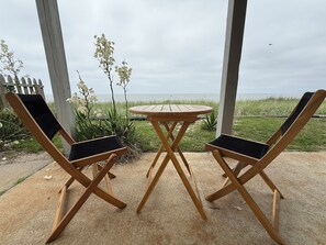 Ett träbord och två stolar med svarta kuddar, placerade på en betongterrass med utsikt över havet och en gräsyta.