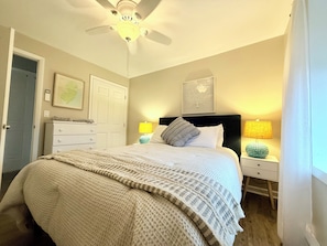 Un dormitorio con cama, ventilador de techo, mesita de noche con lámpara, cómoda y una puerta.