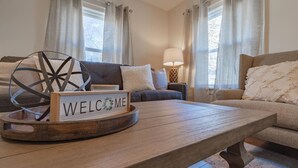 Ein gemütliches Wohnzimmer mit einem Holztisch, einem 'WELCOME'-Schild, einer dekorativen Metallkugel und einer Couch mit Kissen.