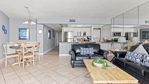 A modern living room with a dining area, a black leather sofa, a wooden coffee table, and a kitchen in the background. - Hello Sunshine Edgewater, Tower 1, #905-Beachfront! 2Bd-2Ba-Sleeps 6-Fun In The Sun! (Panama City Beach)